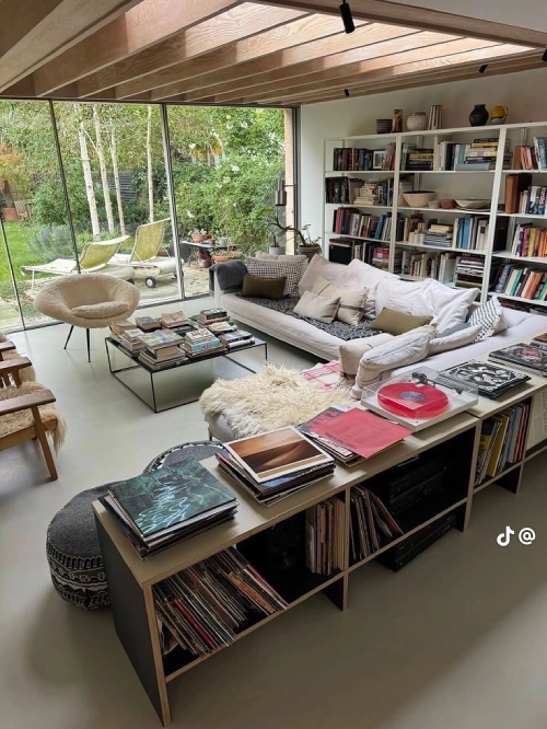 Someone's living room with shelving filled with books and storage units around the sofa filled with vinyl records. There are books on the glass coffee table in the centre and the patio windows go out to the garden. It has a modernist vibe.