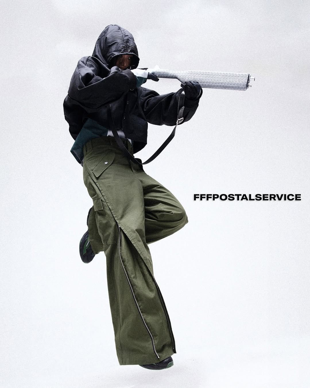 a person in a hooded jacket and long baggy khaki trousers, pointing a gun-type weapon covered in a knitted cover