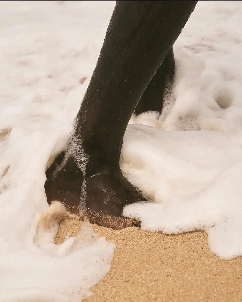 A Black person's foot in the sand with the sea water hitting it