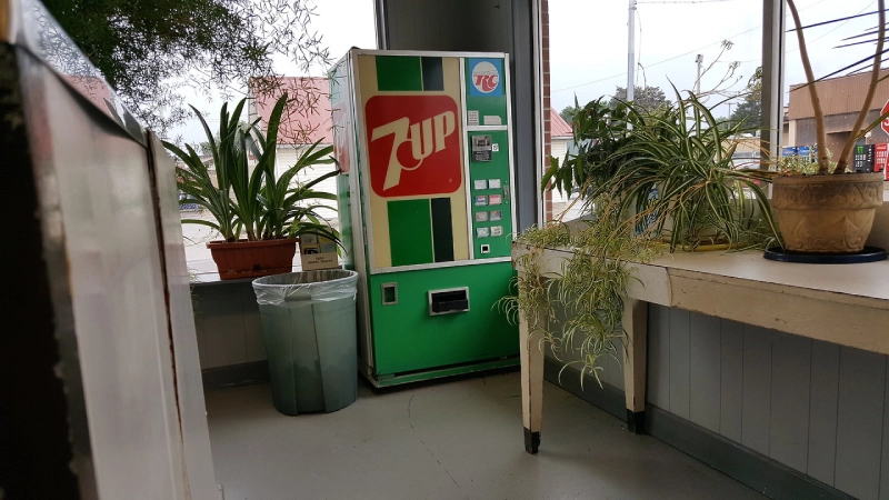 a 7up vending machine in a laundrette with indoor plants around it and a bin next to it